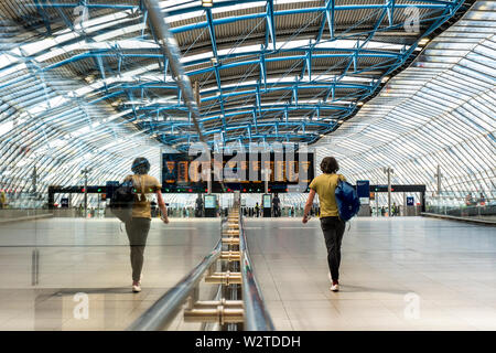 WATERLOO STATION single lone rail traveller in quiet new architecture surrounding platform additions 20-24 in former Eurostar Terminal. Modern busy departures concourse with single  rail traveller walking to platforms in late afternoon sun reflected in glass barrier, network information screens in background Waterloo Station London SE1 Stock Photo