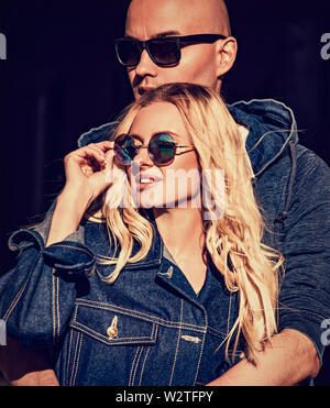 Stilish couple cuddling in modern blue jeans jackets and trendy sunglasses on summer city background. Beautiful female blond model. Closeup vintage to Stock Photo