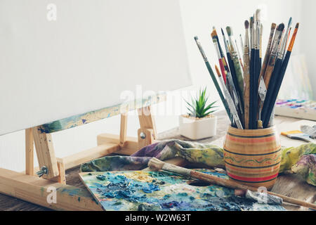 Artistic equipment in studio: canvas on wooden easel, paint brushes, paints and used palette.  Copy space for text. Stock Photo