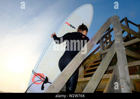 A young woman carries her surfboard up the steep wooden steps at Compton Chine on the Isle of Wight Stock Photo