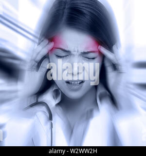 Young woman with a pounding headache or migraine standing clutching her temples with an expression of pain, monochrome image with selective red color to temples and blur effect around her face Stock Photo