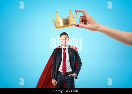 Female hand holding golden crown above young businessman with red cloak on blue background Stock Photo