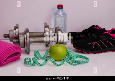 Female fitness. Green apple with dumbbells, a bottle of water, a rug, running shoes and a tape for measuring volumes on a white background. Stock Photo