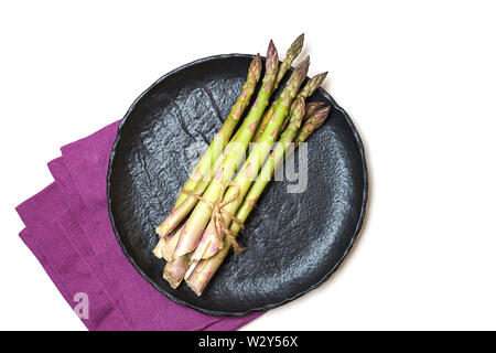 Fresh green asparagus on black ceramic dish and pink linen napkin on white background, isolated. Stock Photo