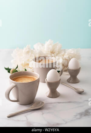Boiled eggs in ceramic egg cups, two cups of coffee with ceramic spoons on background of beautiful white peony. Breakfast concept. Copy space. Stock Photo
