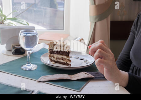 A slice of multi-layered walnut cake with almond nut plates on a chocolate-decorated plate on a table decorated with napkins in a cafe. An old woman h Stock Photo