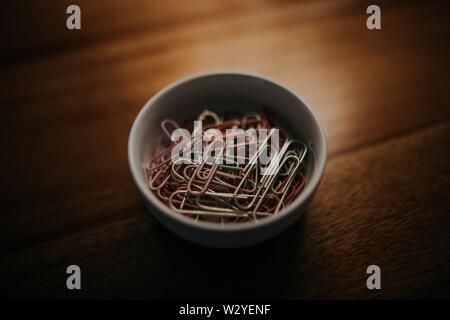Pile of paper clips in a ceramic bowl on a brown wooden table Stock Photo