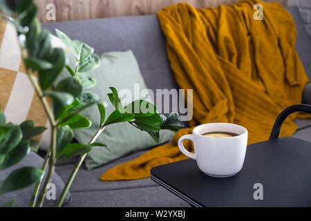 A cup of hot tea in a white cup on the table near a cozy gray sofa with pillows and a blanket. Home Autumn Lifestyle Stock Photo