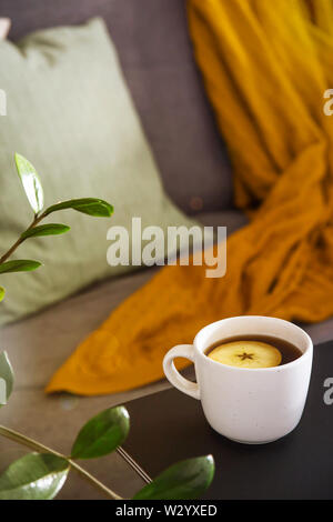 A cup of hot tea in a white cup on the table near a cozy gray sofa with pillows and a blanket. Home Autumn Lifestyle Stock Photo