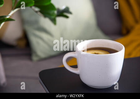 A cup of hot tea in a white cup on the table near a cozy gray sofa with pillows and a blanket. Home Autumn Lifestyle Stock Photo
