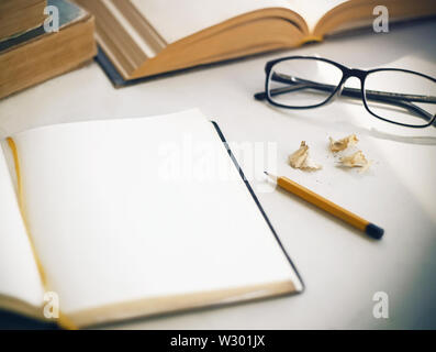 On the white table there are old books, an open diary with a yellow bookmark for notes, shavings from a sharpened, a yellow pencil and glasses in a bl Stock Photo