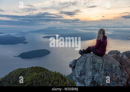 Adventurous Female Hiker on top of a mountain covered in clouds during a vibrant summer sunset. Taken on top of St Mark's Summit, West Vancouver, Brit Stock Photo