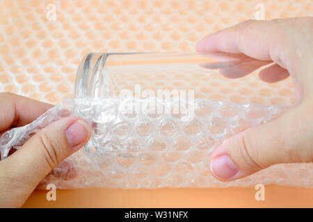 Woman hands package glass for water with transparent bubble wrap on a yellow background. Material for packing fragile items for safe transportation. Stock Photo