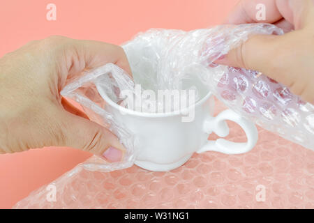 Woman hands package white tea cup with transparent bubble wrap on a pastel pink background. Material for packing fragile items for safe transportation Stock Photo