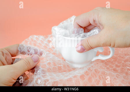 Woman hands packaging a white tea cup with transparent bubble wrap on a pink background. Material for packing fragile items for safe transportation. Stock Photo