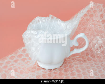 White elegant tea cup half packaged with transparent bubble wrap on a pink background. Material for packing fragile items for safe transportation. Stock Photo