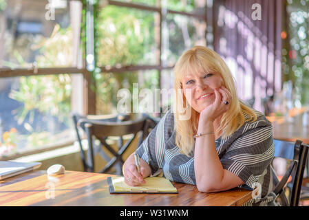Portrait of beautiful plus size attractive senior woman smiling and writing diary in cafe. Stock Photo