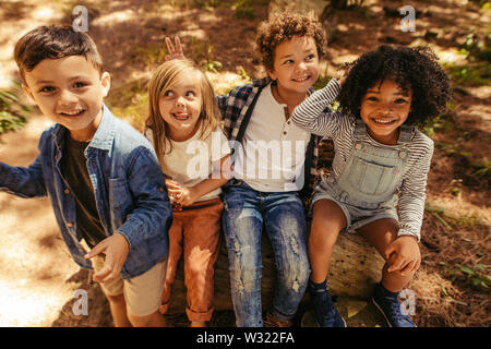 Group of four kids sitting on a wooden log and having fun. Multi-ethnic group of kids playing in a park. Stock Photo