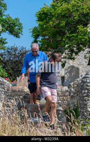 Walkers on the public footpath  using the  Stone steps stile to cross over on the flint  stone wall at St Peter and  St Paul Church, West Wittering Stock Photo