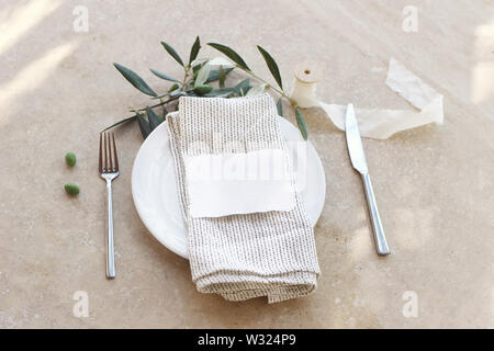 Festive table summer setting with silver cutlery, olive branch, porcelain plate, linen napkin. Place card mockup. Marble background in sunlight Stock Photo