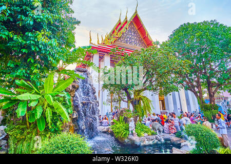 BANGKOK, THAILAND - APRIL 22, 2019: The tourists rest on the shady stone border of the waterfall's pond and prepare for further exploring Wat Pho reli