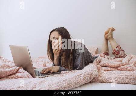 Young adult beautiful girl enjoys a laptop in bed and yawns. She was tired or woke up early and sleepy. Stock Photo