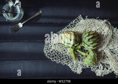 Top view Fresh green artichokes with braided rope and kitchen stuff on the black wooden table lightened by morning window light. Farmer harvest, local Stock Photo