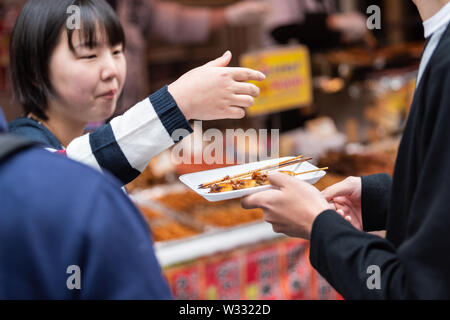 Tokyo, Japan - March 30, 2019: Chinese woman, man people holding skewered scallops on skewer stick on platter or tray in Tsukiji outer outdoor street Stock Photo