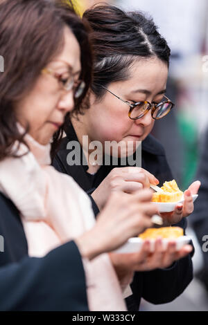 Tokyo, Japan - March 30, 2019: Side view of two Asian Chinese women eating tamagoyaki Japanese eggs rolled omelet in Tsukiji fish street outer market Stock Photo