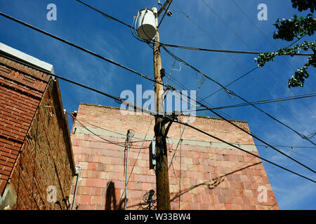 An upward view of several electrical lines and telephone cables intersecting at a wooden light pole against old red brick and concrete buildings, USA Stock Photo