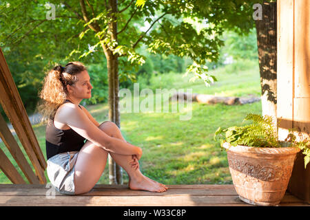 Spring house porch with happy woman sitting on steps of house in front or back yard morning wooden cabin cottage Stock Photo