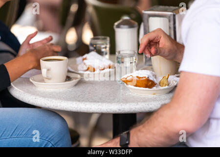 Closeup of couple sitting outside at sidewalk cafe by table drinking chicory coffee and eating deep fried beignet donut powdered with sugar
