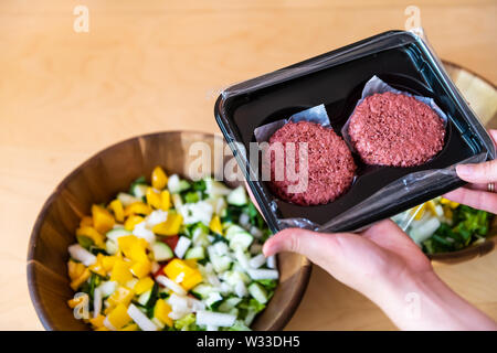 Woman holding two raw uncooked red vegan meat burger patties by salad bowls in packaging Stock Photo