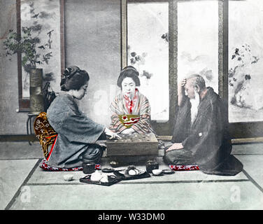 [ 1890s Japan - Playing Go ] —   Two young women in kimono and traditional hairstyles and an elderly man in a haori coat are seated around a board for the game of go.  The studio is arranged as a zashiki (guest room), with a screen, an ikebana flower arrangement and scroll in the back. A small tray with a teapot and two cups can be seen at the front.  19th century vintage glass slide. Stock Photo