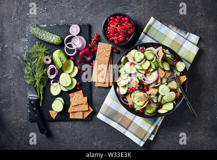 delicious summer salad of cucumber, red currant, marinated red onion and crisp flatbread pieces in a black bowl with ingredients on a concrete table, Stock Photo