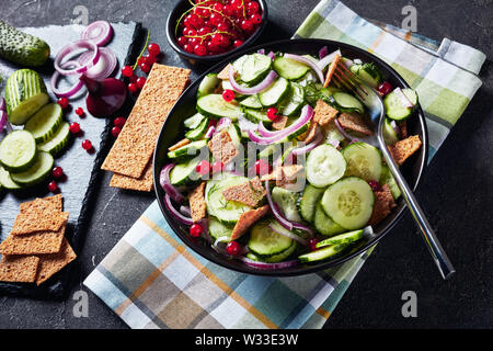 close-up of fresh summer cucumber salad with red currant, marinated red onion and crisp flatbread pieces in a black bowl with ingredients on a slate t Stock Photo