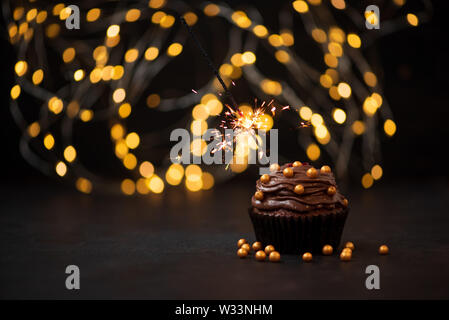 Chocolate cupcake with golden candies and burning sparkler on dark wooden background against blurred lights. Selective focus. Unhealthy food or holida Stock Photo