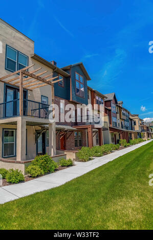 Row of homes with porches and small balconies under blue sky on a sunny day. A paved pathway and grassy terrain can be seen in front of the residences Stock Photo