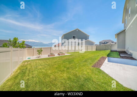 Backyard of a home with small tree and planting beds near the wooden fence. View of houses, lake, mountain, and sky can be seen from this grassy yard. Stock Photo
