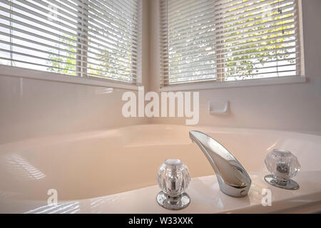 Polished bathtub with shiny faucet and double crystal handle inside a bathroom. Trees and bright sky can be seen through the windows with blinds. Stock Photo