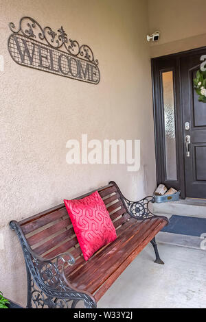 Rustic metal and wood bench with red pillow on the porch of a home. A Welcome sign, security camera, sidelight, and door with wreath can also be seen Stock Photo