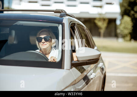 Portrait of a senior woman driver sitting in the modern car, looking out the window Stock Photo