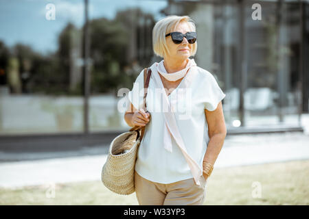 Portrait of a happy and elegant senior woman dressed in white shirt with sunglasses and bag outdoors Stock Photo