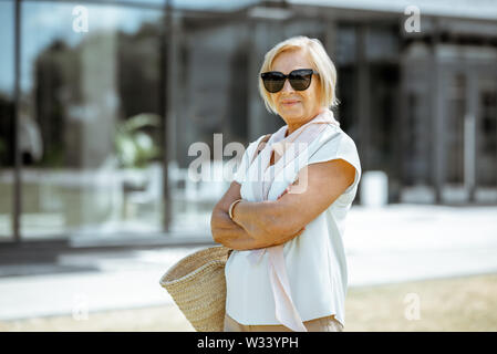 Portrait of a happy and elegant senior woman dressed in white shirt with sunglasses and bag outdoors Stock Photo
