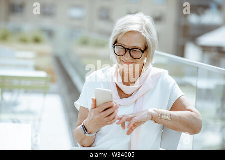 Elegant senior woman using smart phone while sitting on the cafe terrace outdoors in the city. Concept of using modern technologies by elderly people Stock Photo
