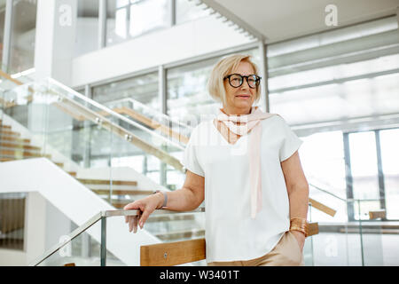 Portrait of a confident senior woman dressed in white shirt standing on the staircase of the modern building Stock Photo