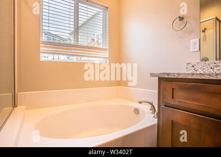 Bathroom interior with oval bathtub against white wall and window with blinds. The vanity has wood cabinet, white countertop with black specks, and mi Stock Photo