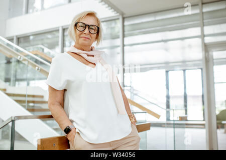 Portrait of a confident senior woman dressed in white shirt standing on the staircase of the modern building