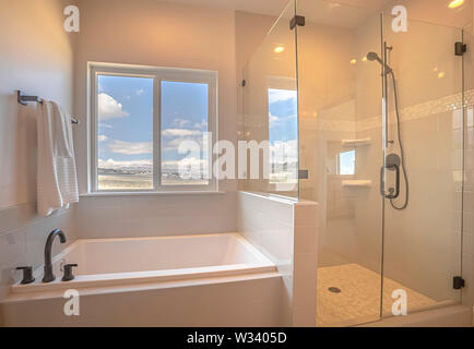 Built in square bathtub inside a bathroom with white wall and sliding window. Inside the shower stall is a tiled floor and wall. Stock Photo