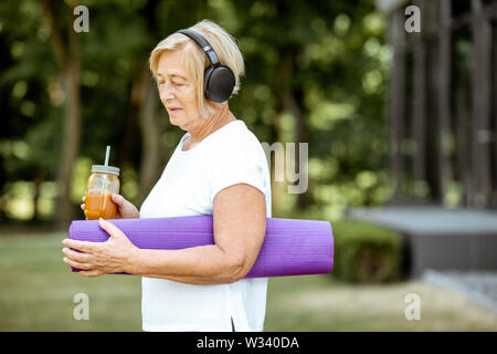 Portrait of an active senior woman in sports clothes standing with yoga mat and juice outdoors. Concept of a healthy lifestyle on retirement Stock Photo